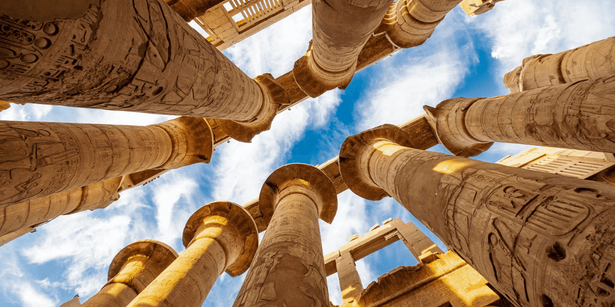 Upward view of ancient Egyptian temple columns with carved hieroglyphs against a blue sky.