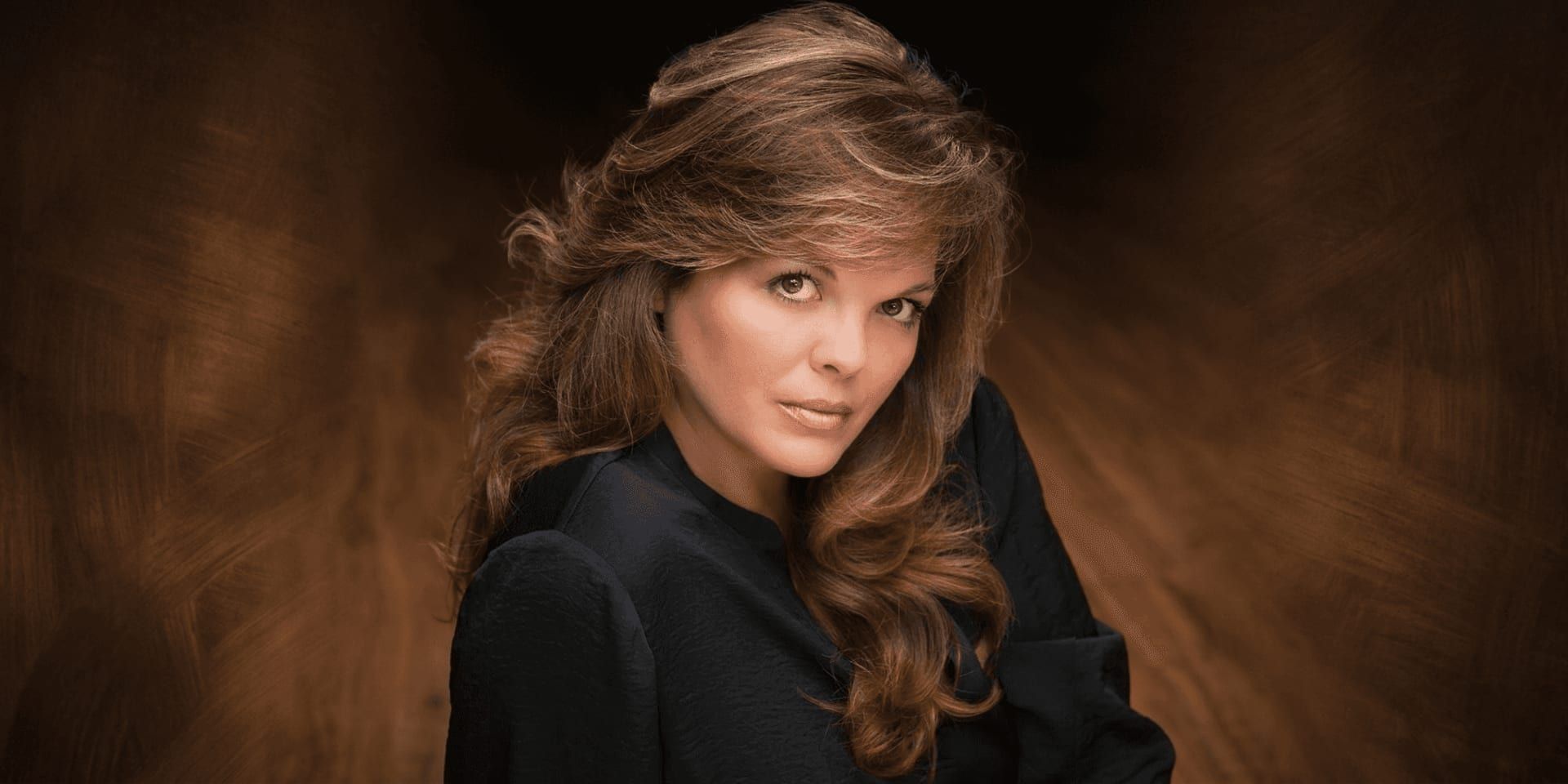 Portrait of a confident woman with voluminous hair, wearing black, against a warm brown studio background.