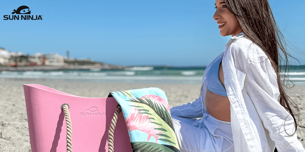 A woman sitting on the beach with a Sun Ninja pink tote bag and a tropical patterned towel, enjoying the warm sunshine and ocean breeze at a scenic beach location.