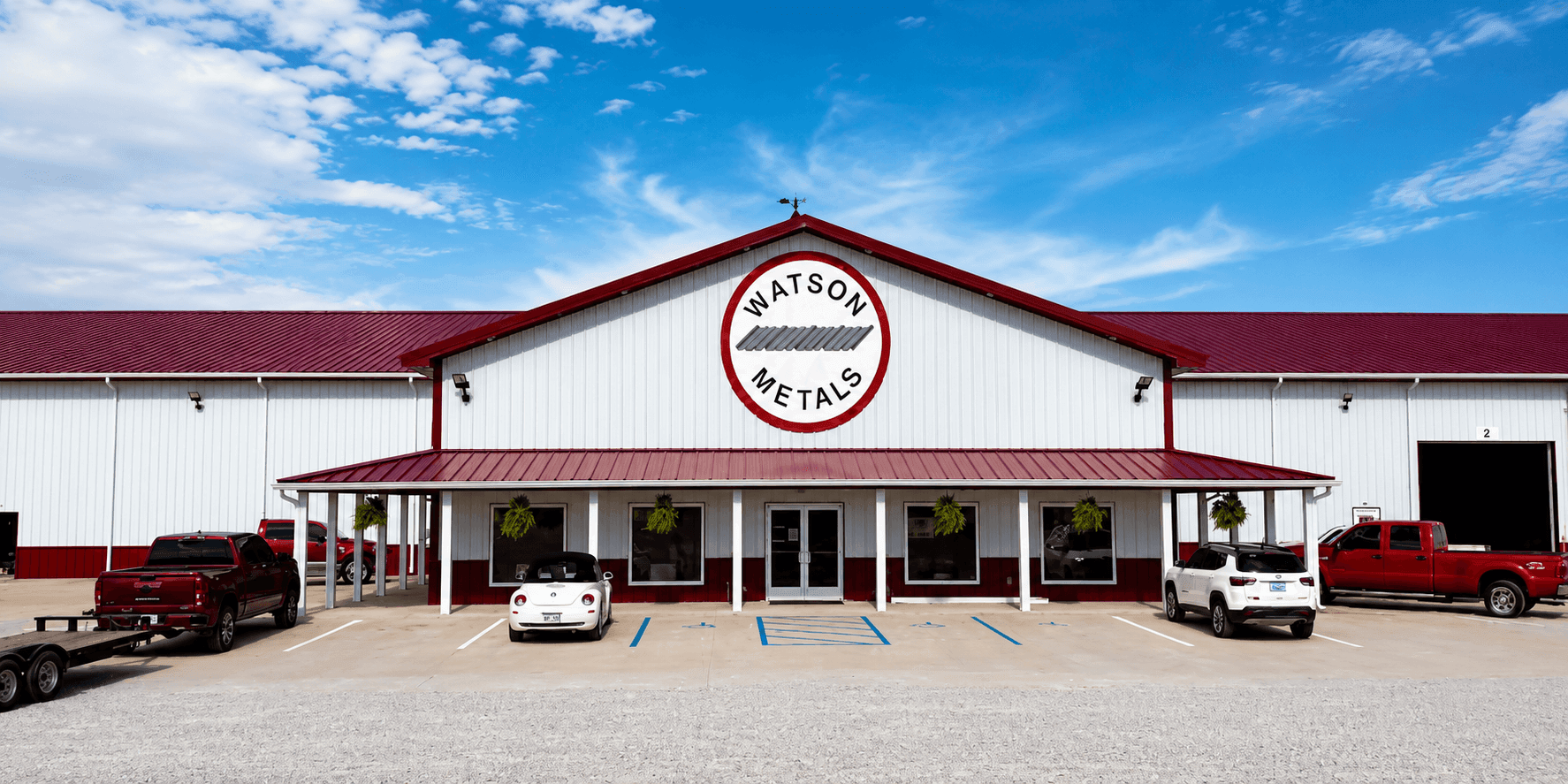 Large white metal commercial building with red roof, Watson Metals logo, covered entrance, and parked vehicles.
