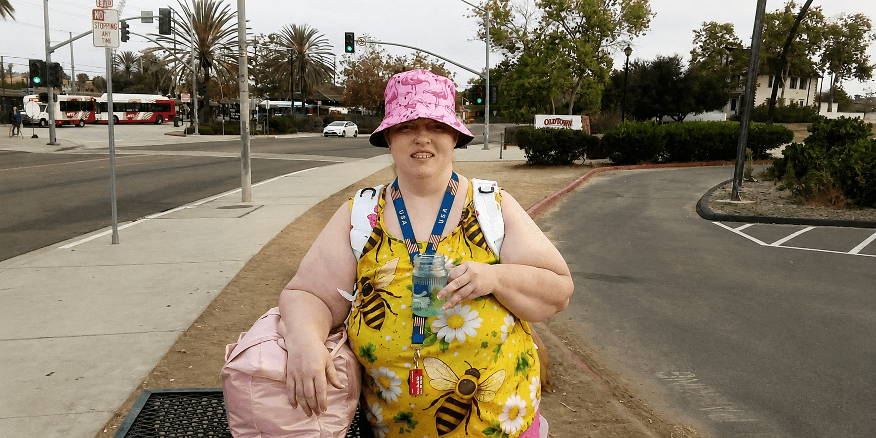 Racheal Ceriani standing outdoors with a backpack, drink, and pink hat near a street intersection.