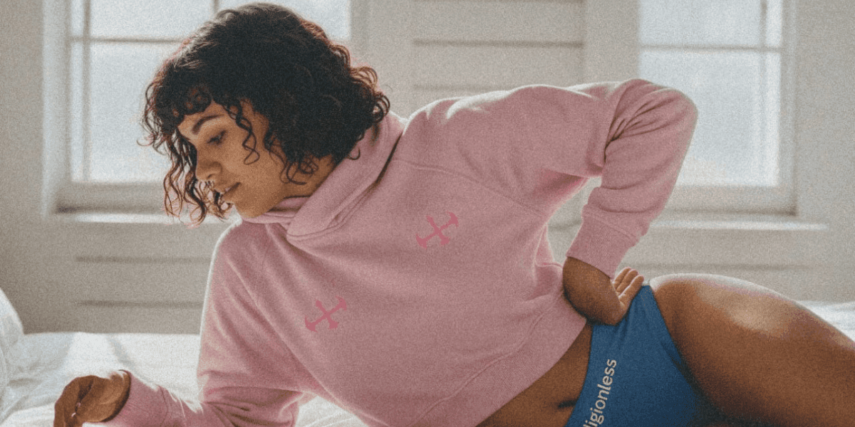 "Woman wearing a pink hoodie with a small logo on the chest, paired with blue shorts, sitting on a bed in a cozy and relaxed indoor setting."