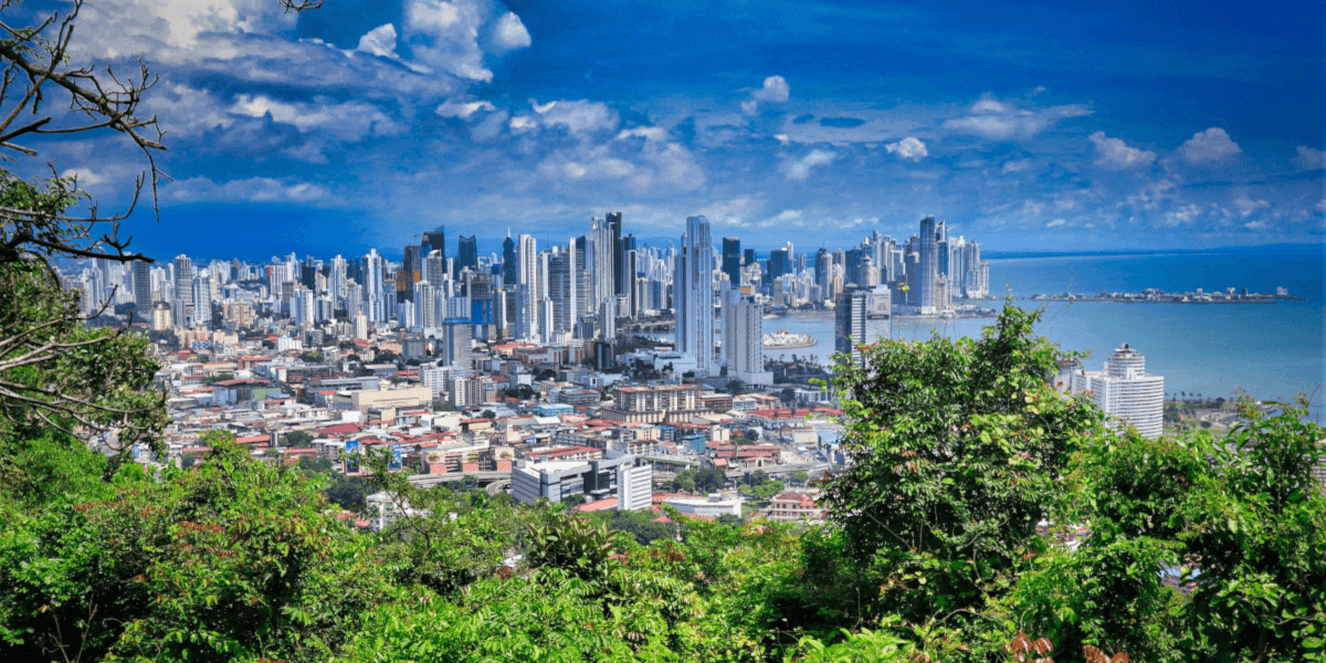 A panoramic view of the city skyline of Panama City, with tall skyscrapers and the coastline visible, surrounded by lush greenery in the foreground under a bright blue sky.