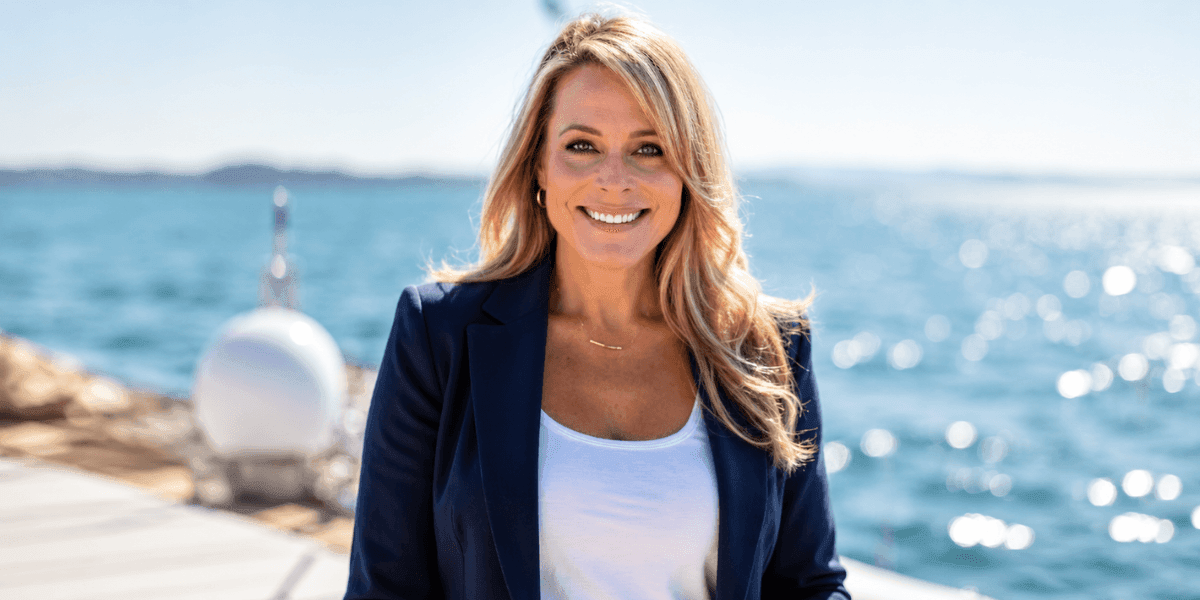 Blonde woman wearing a navy blazer and white top standing outdoors beside the ocean.