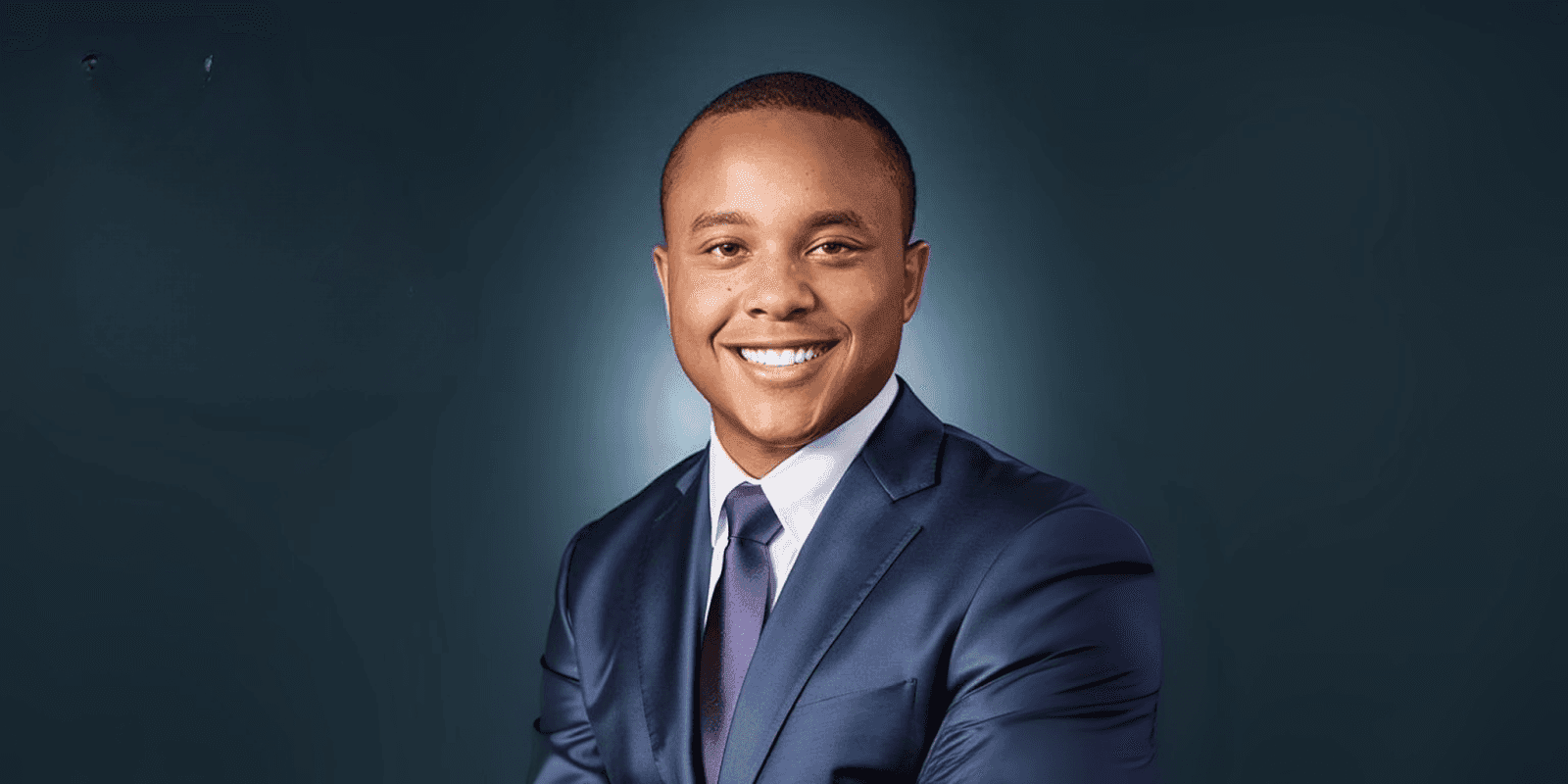 "Close-up of a young professional man wearing a navy suit, smiling with a confident expression against a dark backdrop."