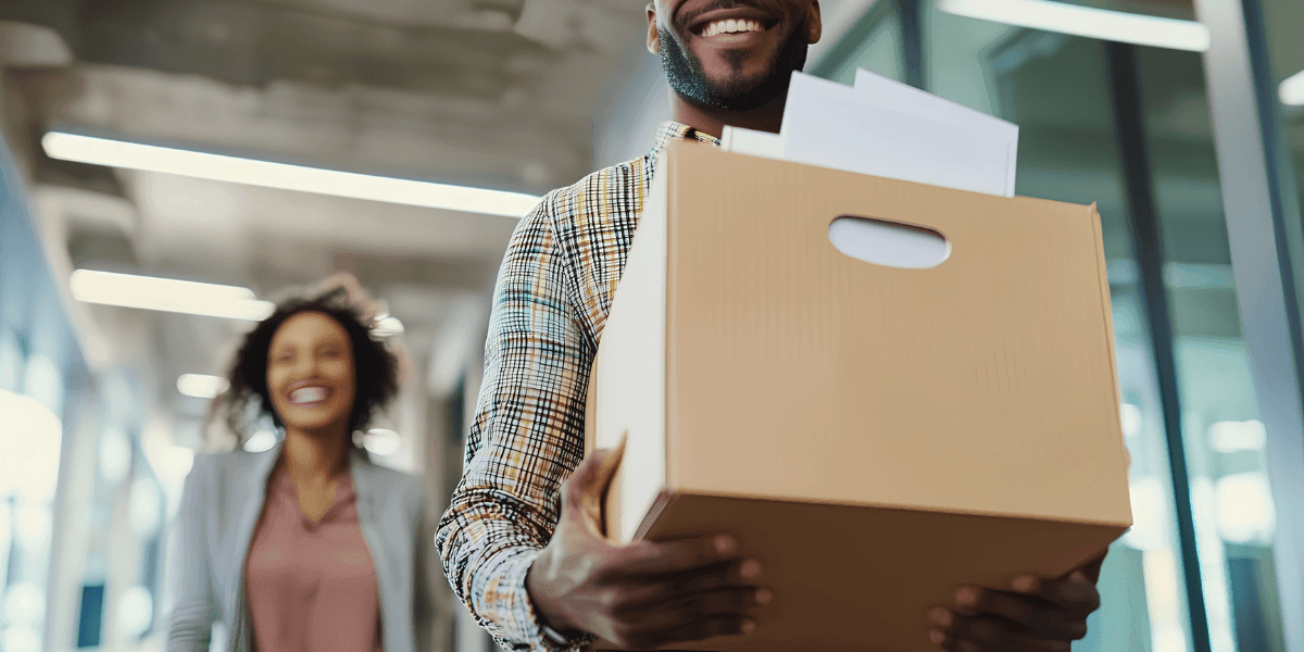 Smiling employee carrying office relocation box while colleague walks through modern workplace corridor.