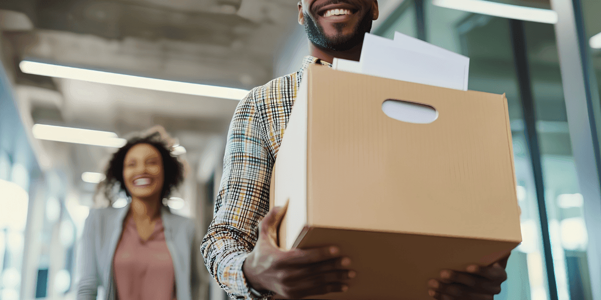 Smiling employee carrying office relocation box while colleague walks through modern workplace corridor.