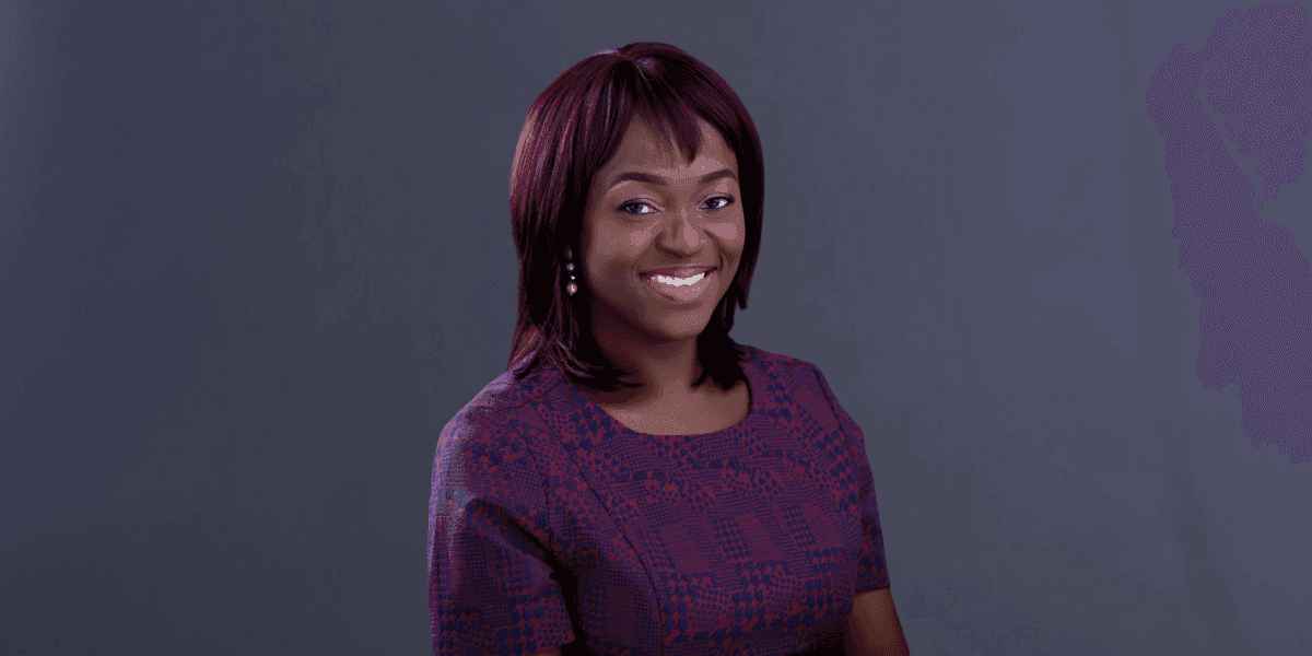 Smiling woman with shoulder-length hair wearing a purple patterned dress against a plain dark background.