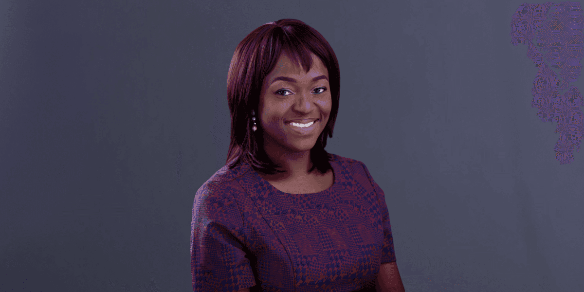 Smiling woman with shoulder-length hair wearing a purple patterned dress against a plain dark background.