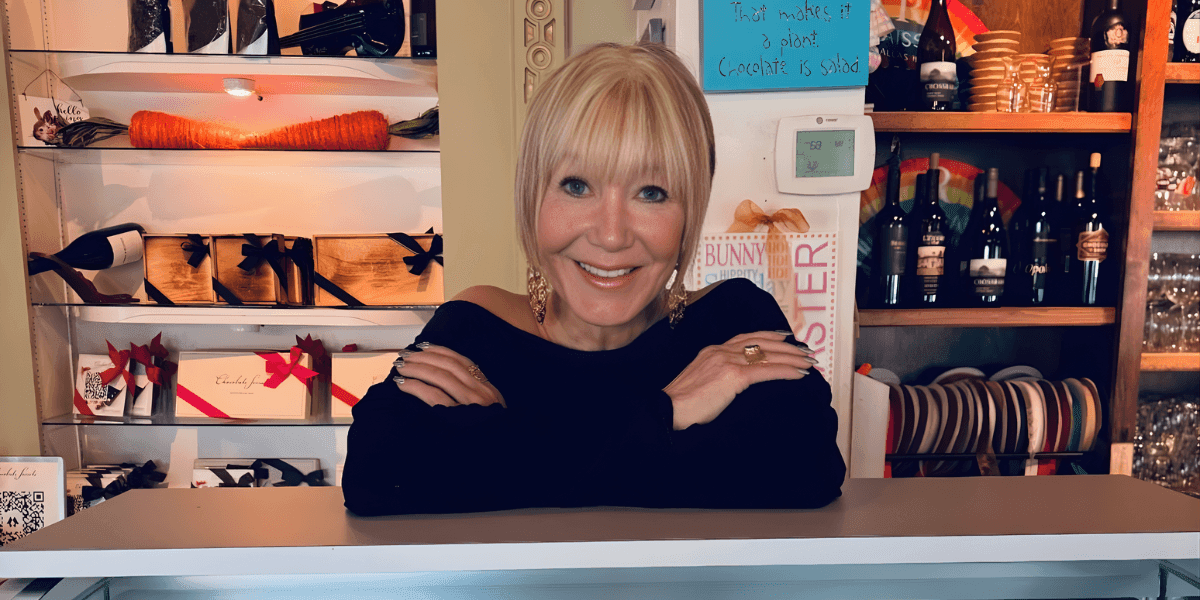 The image shows Suzanne Palmlund, the 88-year-old founder of Enjoy Fun Fudge, smiling behind her store counter, showcasing her business.