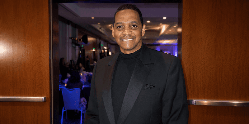 Man in a black tuxedo and turtleneck smiling at an indoor formal event entrance.