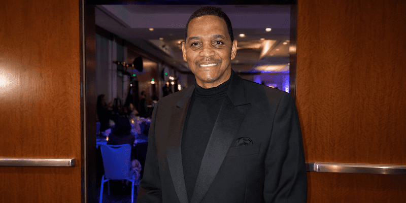 Man in a black tuxedo and turtleneck smiling at an indoor formal event entrance.