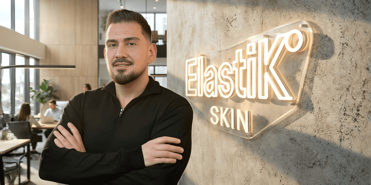 Man with arms crossed in modern office beside illuminated ElastiK Skin sign, coworkers blurred in background.