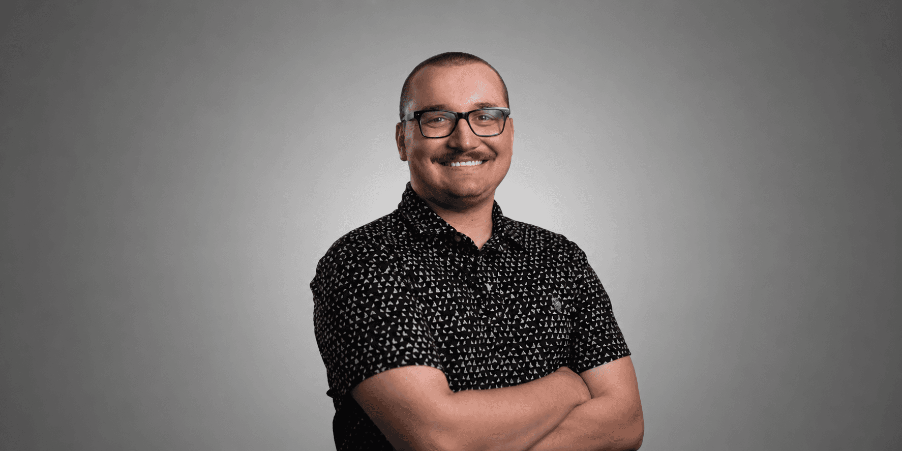 Preston Vawdrey wearing glasses and a patterned black shirt with arms crossed against a gray background.