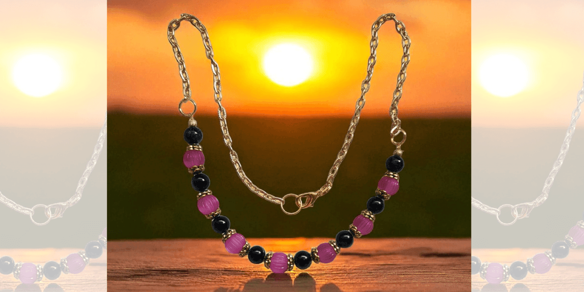 A handcrafted necklace from Crystal’s Beadery featuring black and purple beads with golden accents against a sunset backdrop.