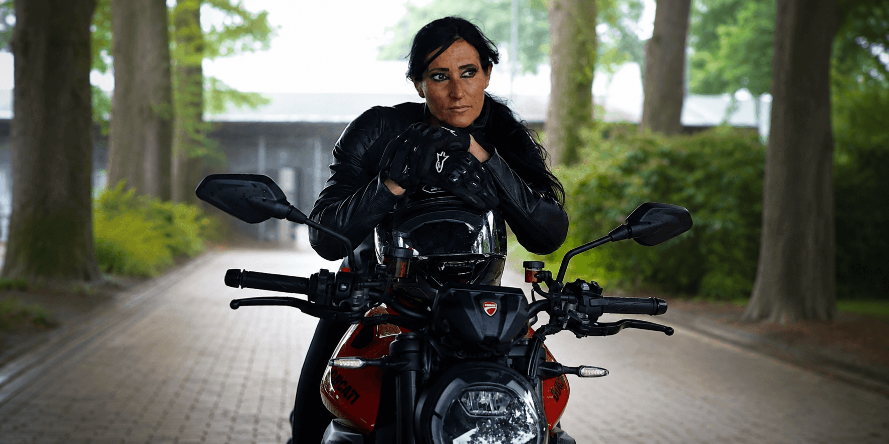 Silvia Rizzo leans on her Ducati motorcycle wearing black riding gear beneath a tree-lined avenue.