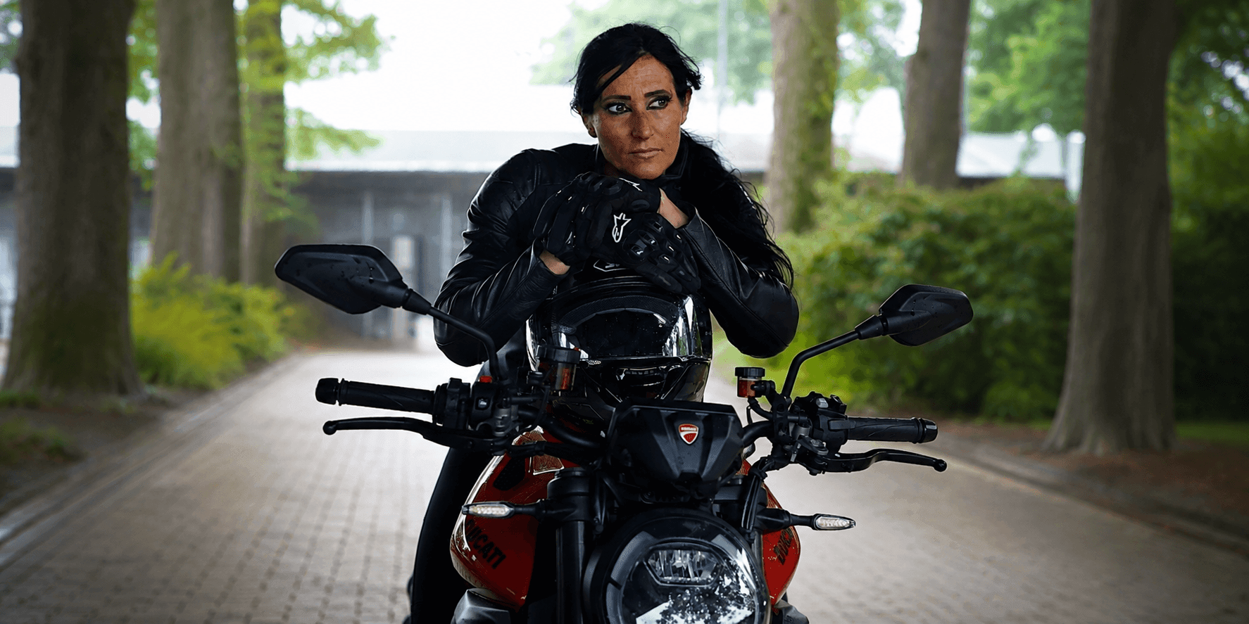 Silvia Rizzo leans on her Ducati motorcycle wearing black riding gear beneath a tree-lined avenue.