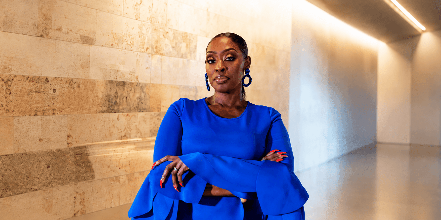 Portrait of Dilys Sillah wearing a blue dress with folded arms in a modern hallway.