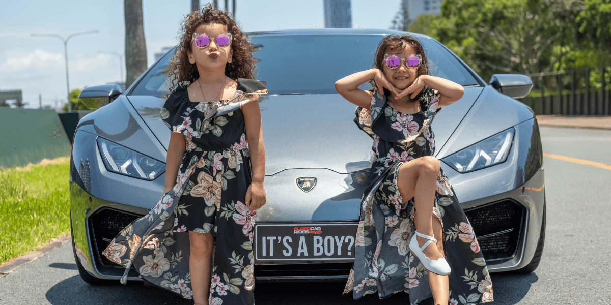 Two young girls in matching floral dresses and sunglasses pose confidently in front of a sleek silver sports car with a "It's a Boy?" license plate.