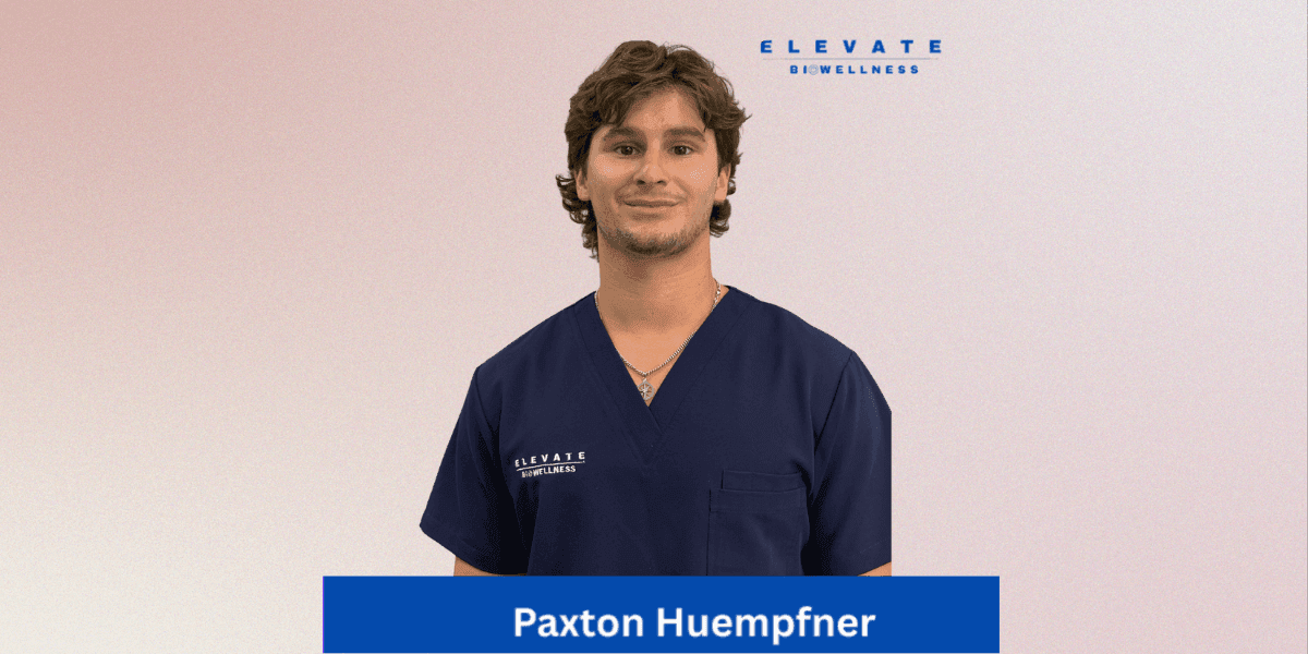 "Portrait of Paxton Huempfner wearing a navy blue scrub top with the 'ELEVATE Biowellness' logo, smiling confidently against a soft pink background."