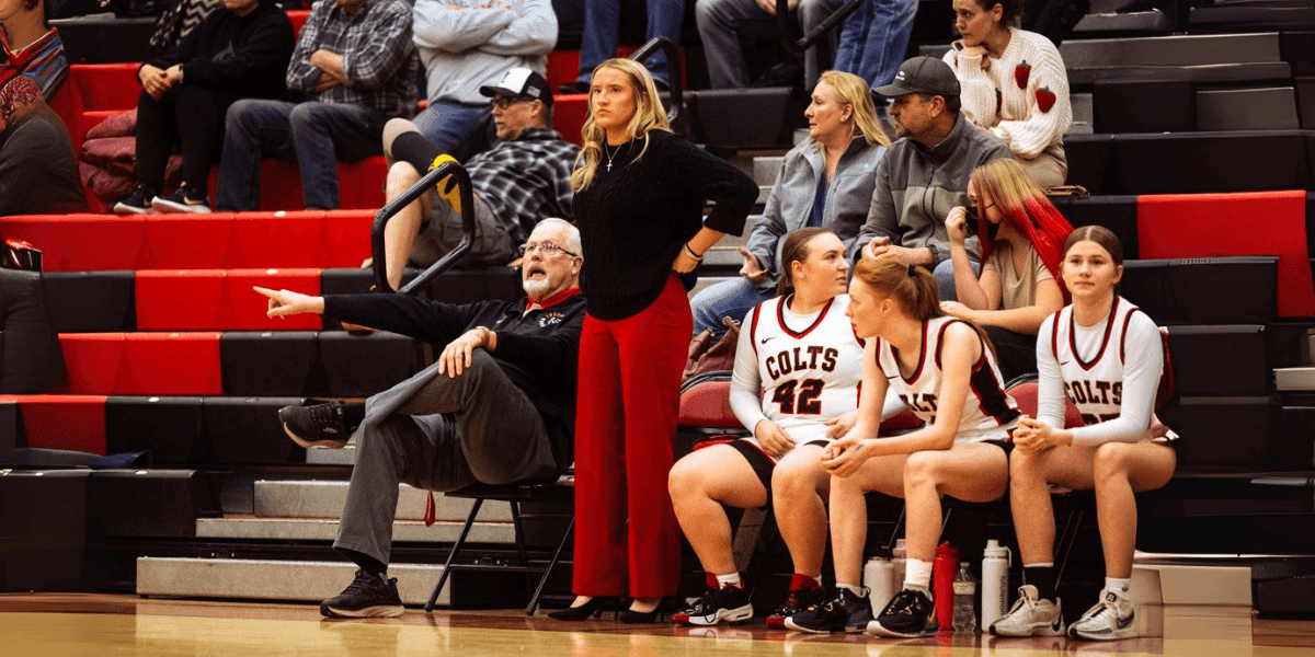 Young female coach stands courtside with players and staff during high school basketball game, symbolizing rising leadership.