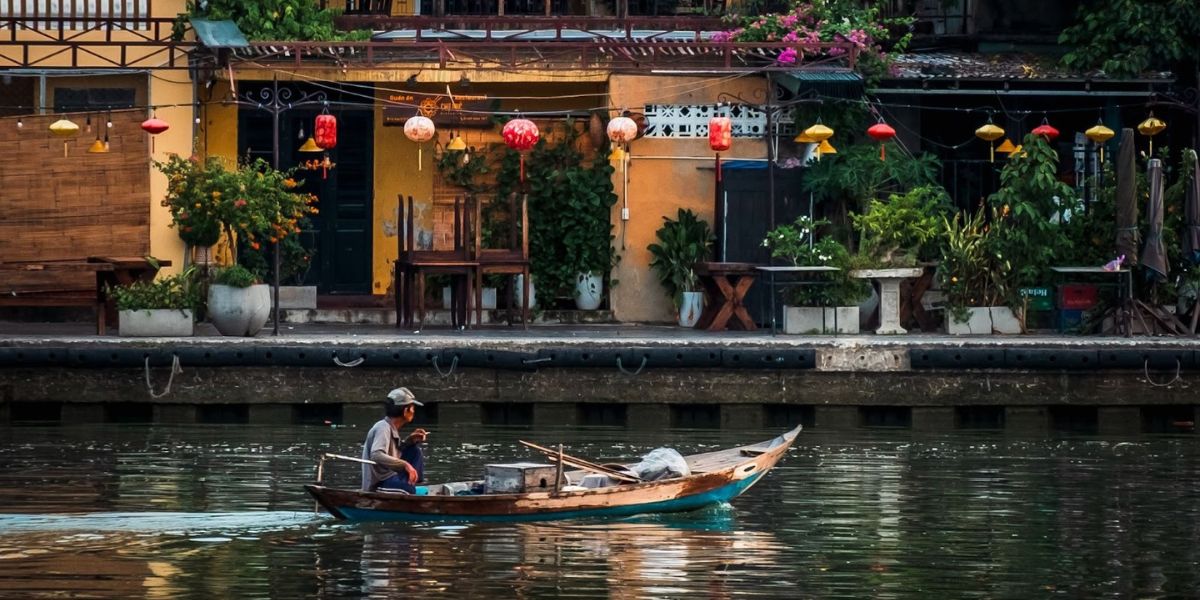 Fisherman on a boat in a Vietnamese waterway, surrounded by colorful lanterns and lush greenery in the background.