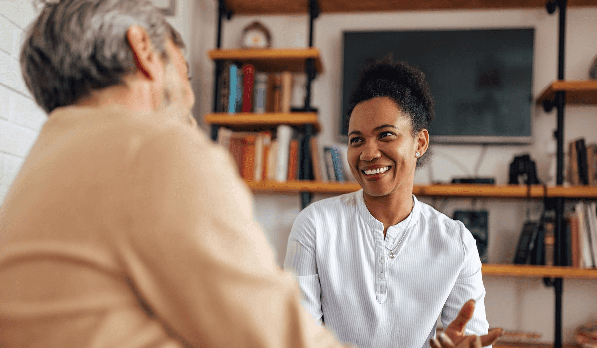 "A woman smiling and engaging in a conversation with an older man. The scene takes place in a cozy indoor setting with bookshelves in the background, creating a warm and inviting atmosphere."