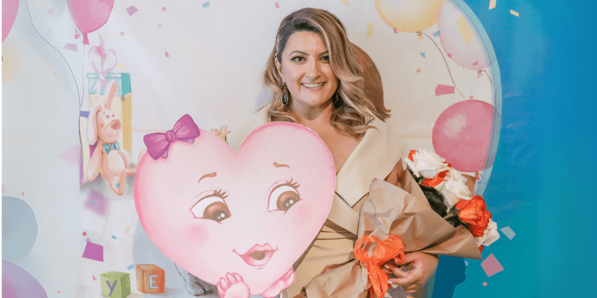 "A woman holding a large heart-shaped prop and a bouquet of roses, standing in front of a colorful backdrop with balloons and confetti. She smiles warmly, dressed in an elegant beige outfit, at a joyful celebration event."