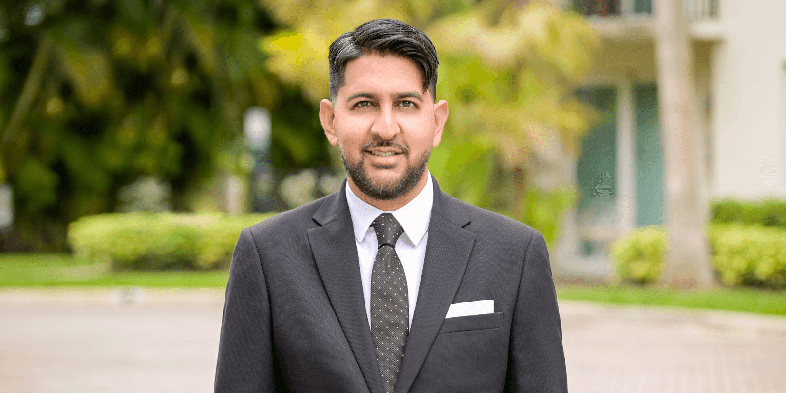 Andrew Roopchand, a luxury real estate agent, confidently poses outdoors in a dark suit, white shirt, and tie.