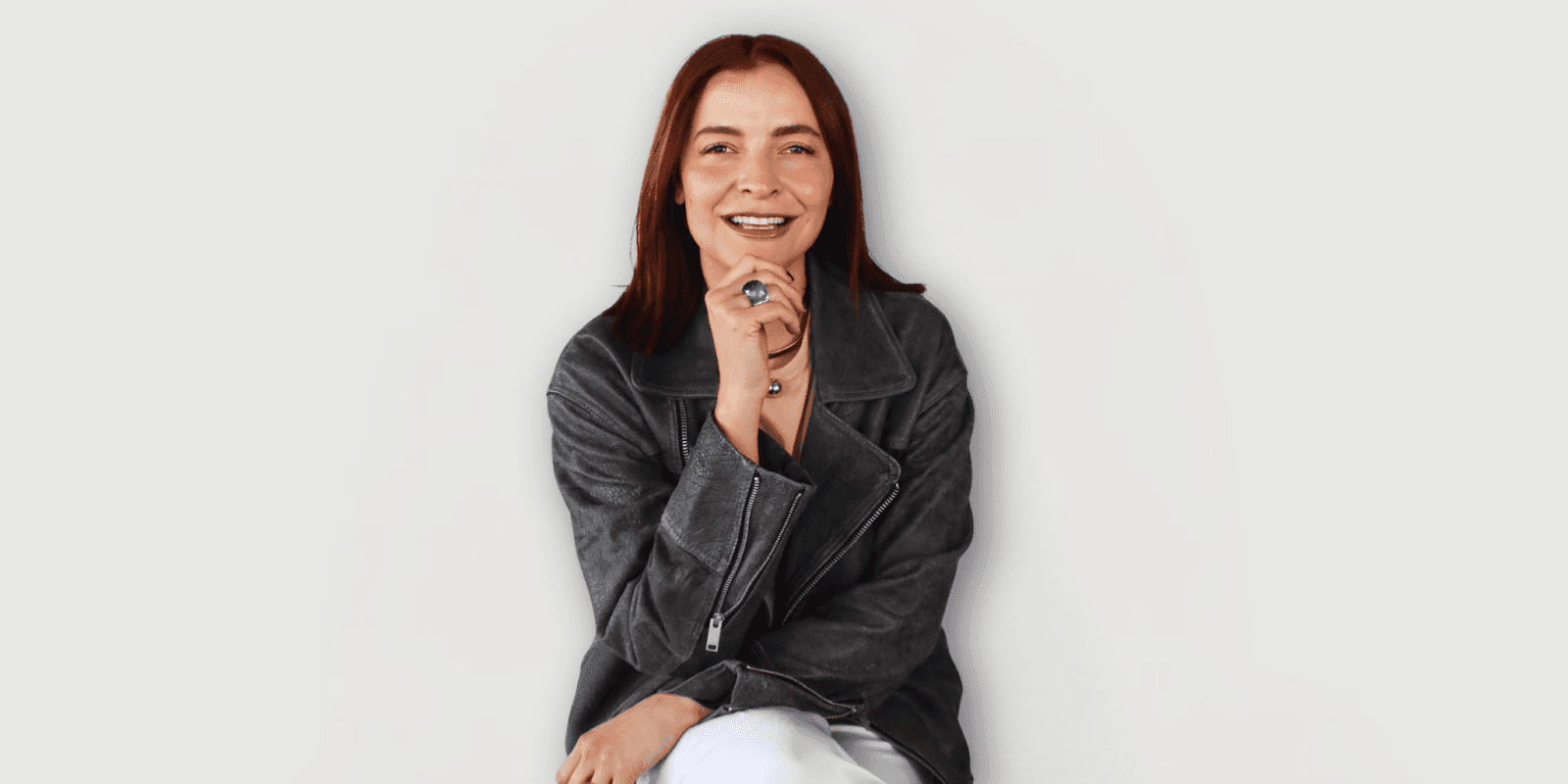 Portrait of a woman with red hair wearing a grey leather jacket, sitting and smiling confidently while resting her hand on her chin against a plain white background.