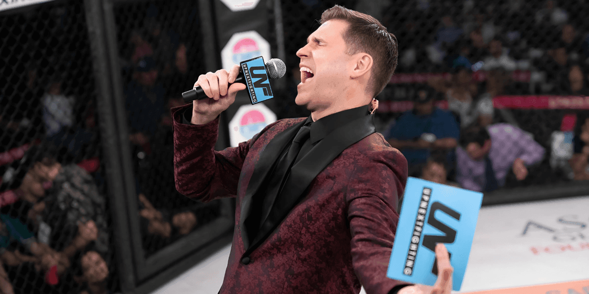 A ring announcer in a maroon suit passionately speaks into a microphone at a live fighting event, with a cheering crowd.