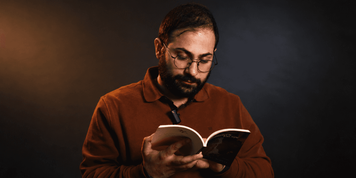 Johnny Achkar wearing glasses and reading A Prisoner’s Escape against a dark studio background.