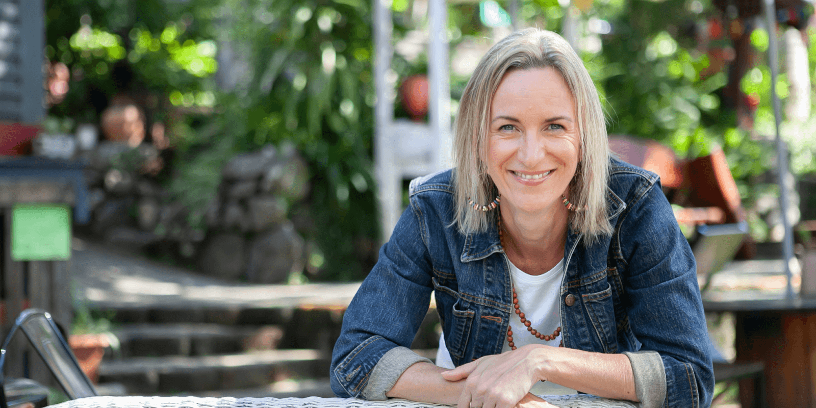 Smiling woman in denim jacket leaning on outdoor table with greenery in the background.