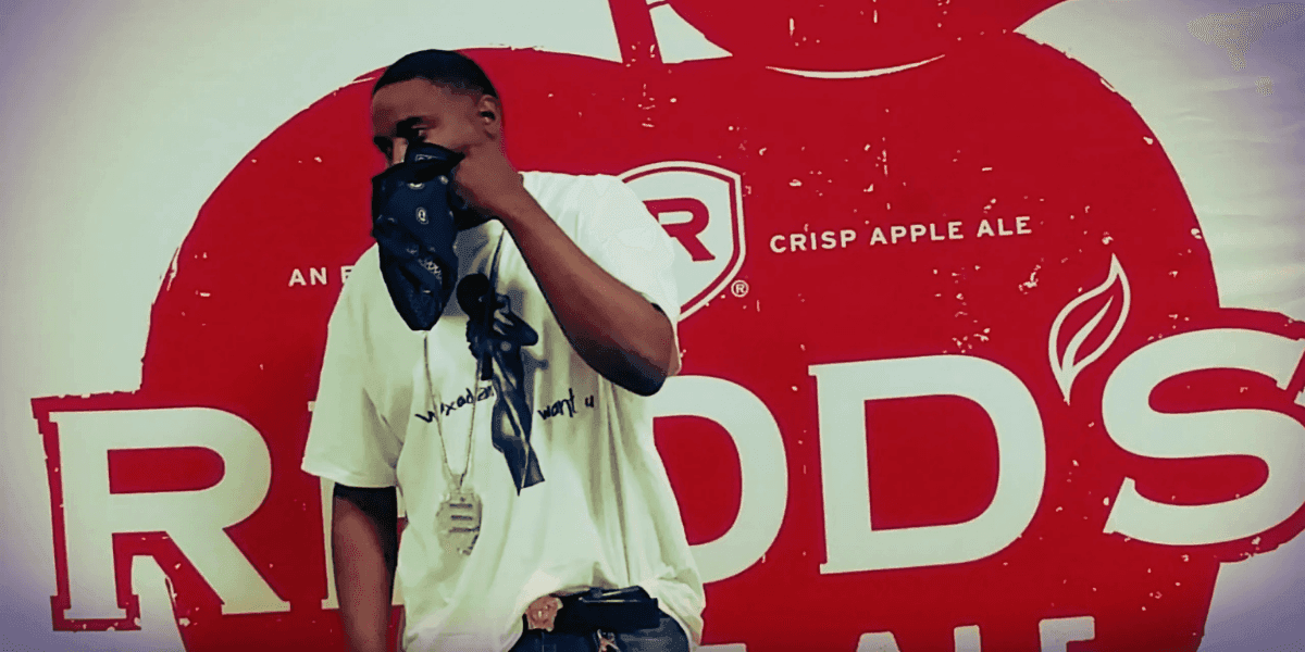 Person wearing a white T-shirt with text, a necklace, and a bandana covering their face, standing in front of a red background with the Redd's Crisp Apple Ale logo.