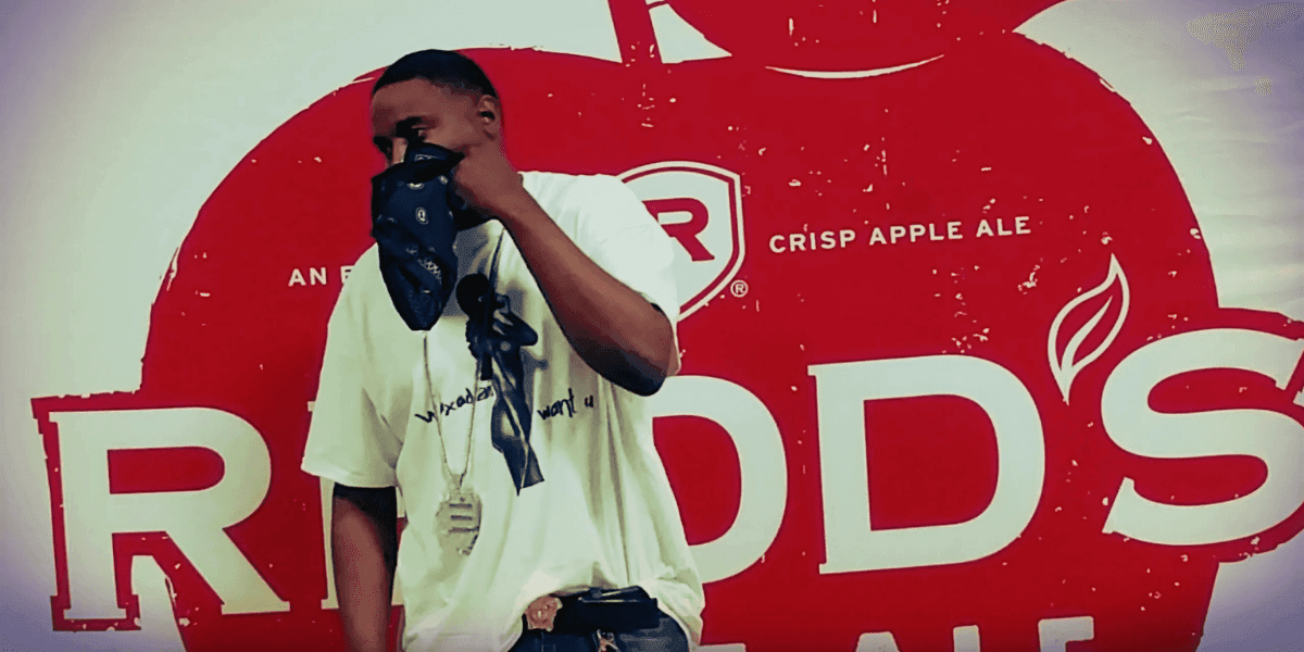 Person wearing a white T-shirt with text, a necklace, and a bandana covering their face, standing in front of a red background with the Redd's Crisp Apple Ale logo.