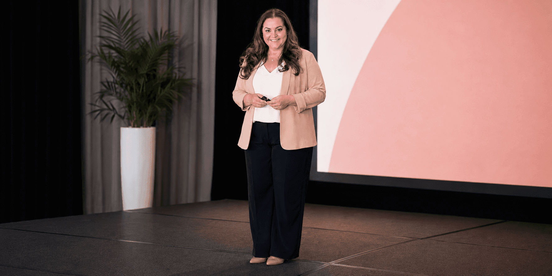 Jackie Cook stands onstage holding a presentation remote beside a large screen.