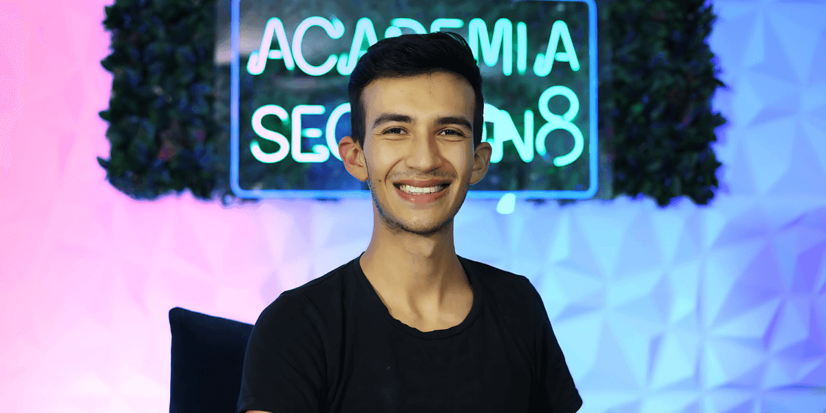 Smiling young man posing in front of a neon sign that reads 'Academia Secreta.' He is wearing a black t-shirt, with a backdrop of greenery and geometric wall design, giving a friendly and approachable vibe.