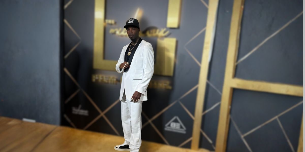 Amazin Jackson stands confidently in a white suit with a black shirt, accessorized with gold jewelry, black sneakers, and a cap.