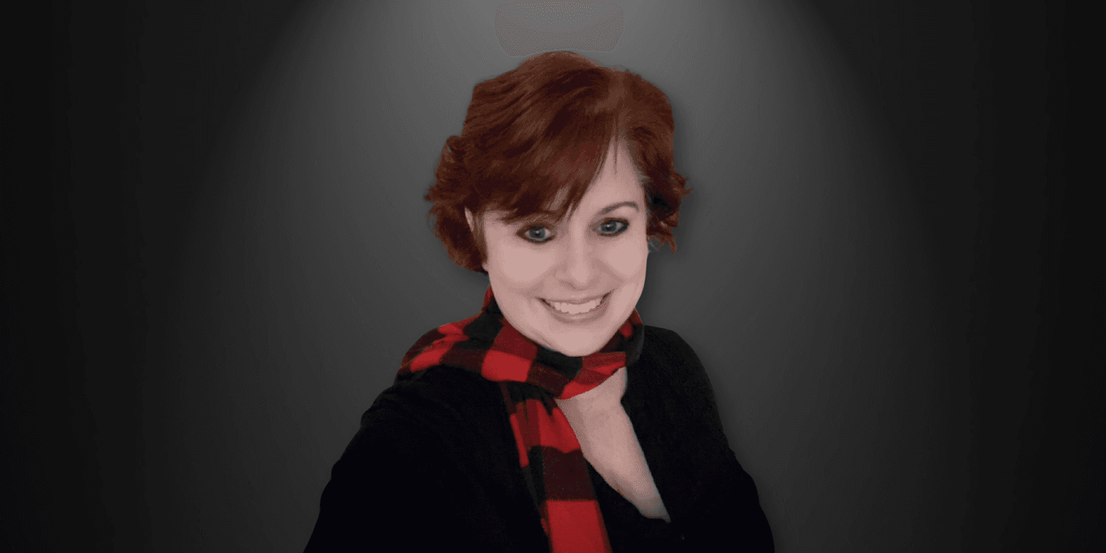 Portrait of a smiling woman with short red hair, wearing a black shirt and red checkered scarf, against a dark background.