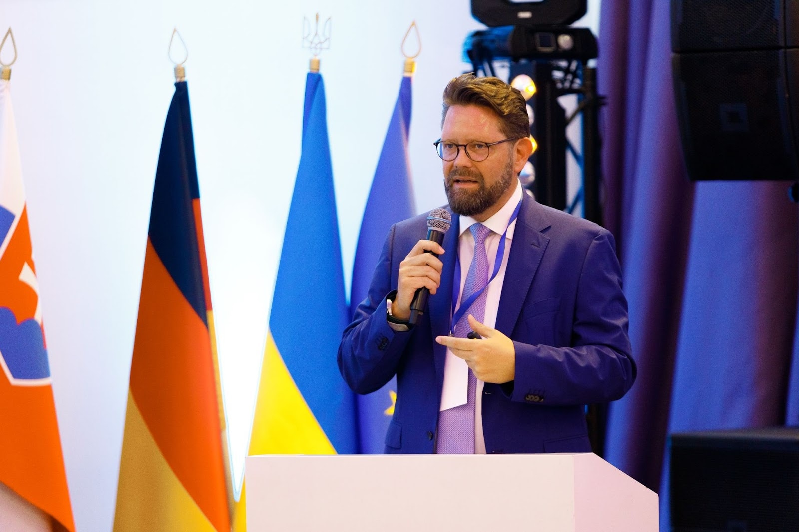 Image of a man in a blue suit and glasses, speaking into a microphone at a conference with flags of Ukraine, Germany, and other European nations in the background.
