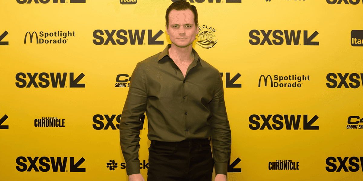 Aaron Dalla Villa poses on the SXSW red carpet in a dark green shirt against a yellow backdrop.