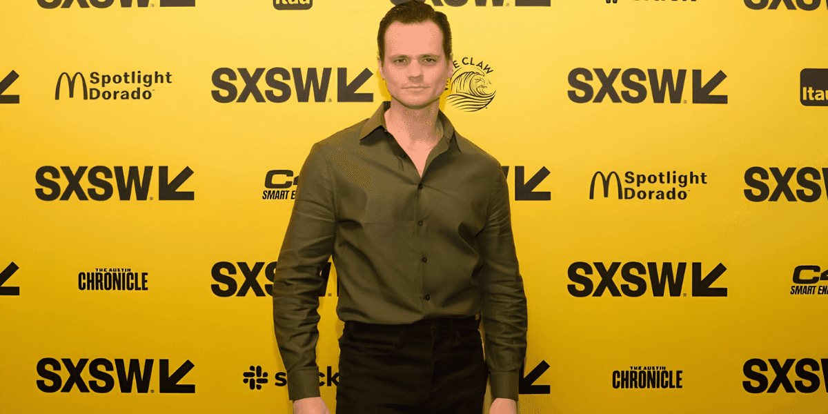 Aaron Dalla Villa poses on the SXSW red carpet in a dark green shirt against a yellow backdrop.