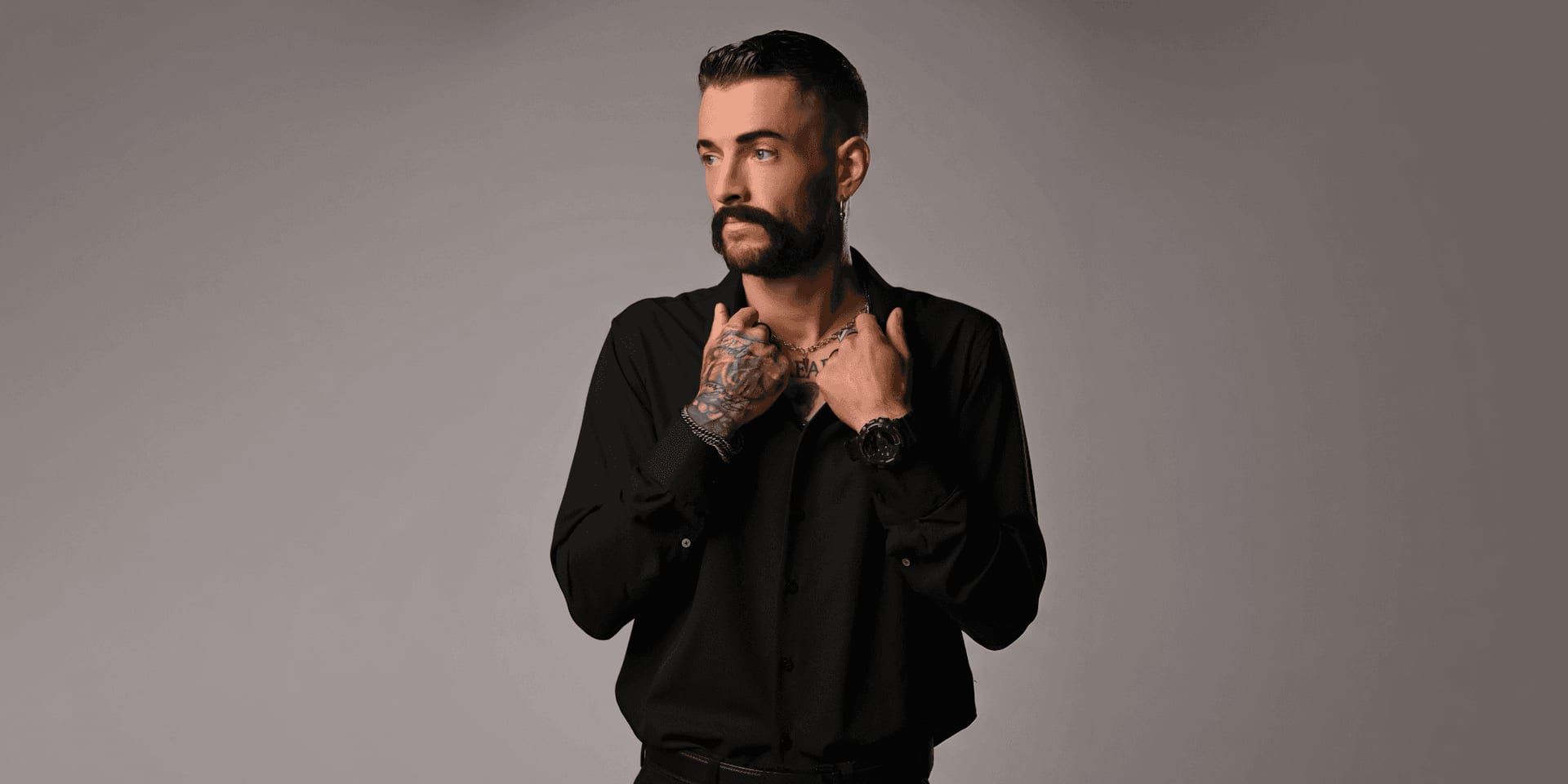 Tattooed man in black button-up shirt posing against neutral studio backdrop, looking to the side.