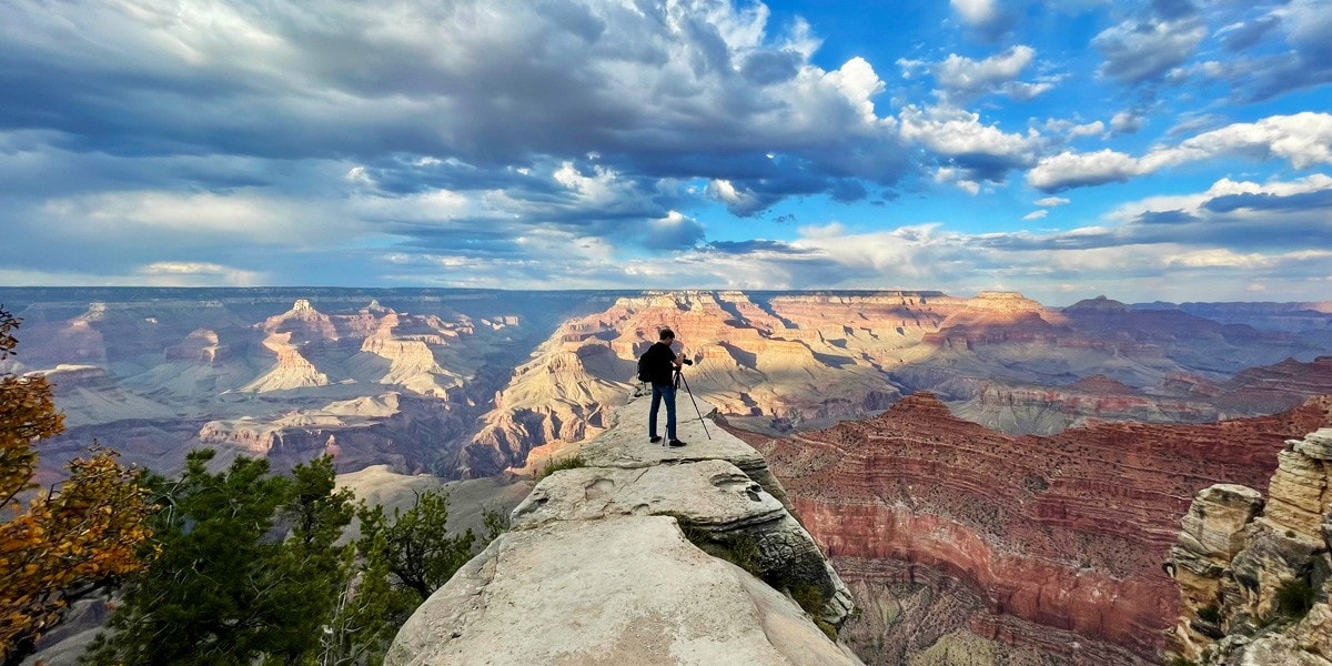 A photographer standing on a narrow ledge at the Grand Canyon, capturing the breathtaking view of the canyon’s deep cliffs and expansive sky