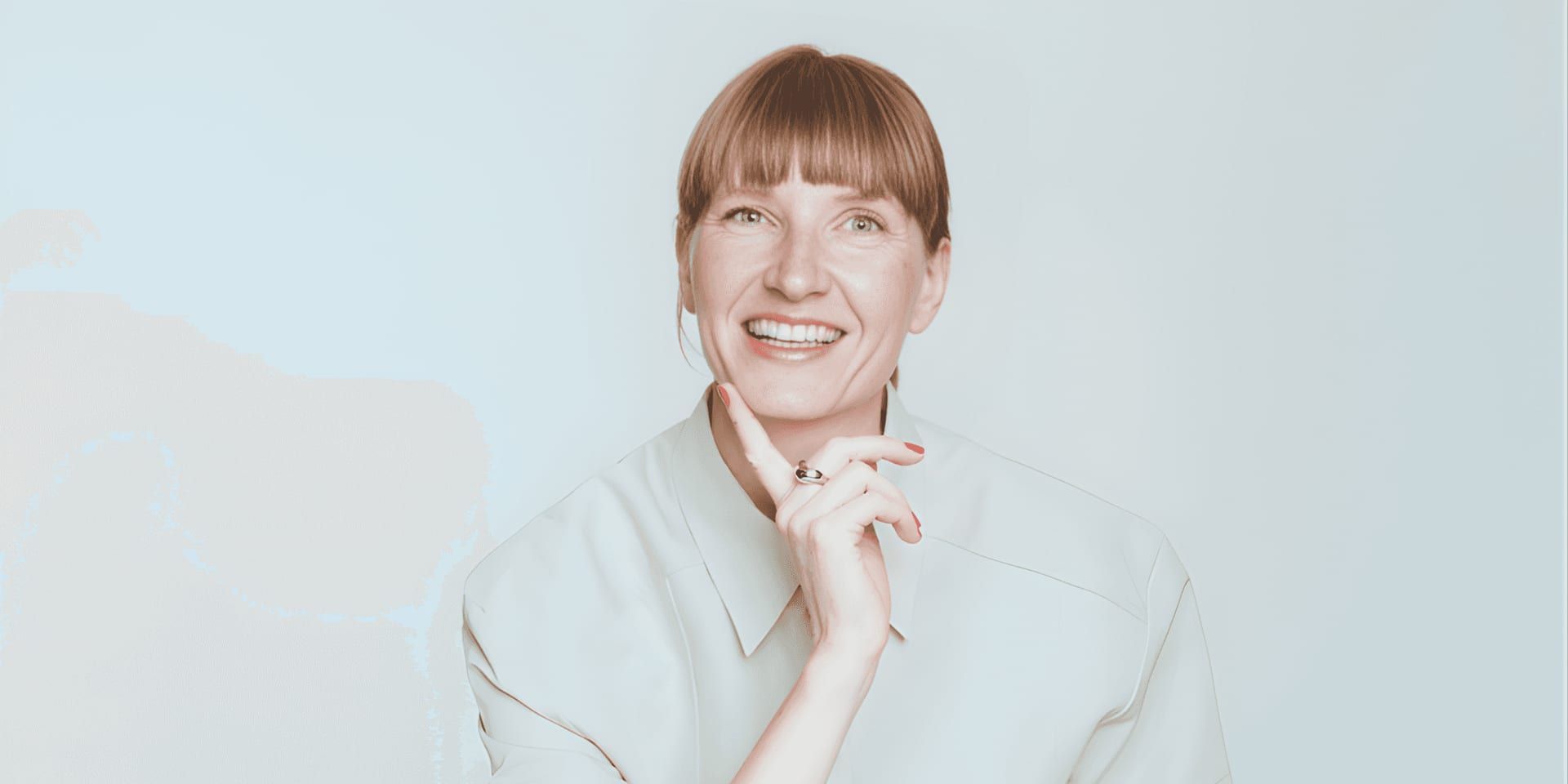 A person with a smiling expression, light brown hair, and bangs poses against a plain background in a light beige shirt, exuding a cheerful and confident tone.