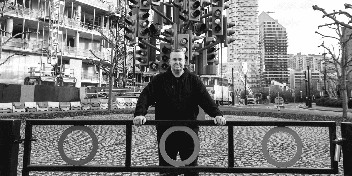 A man stands confidently in front of a sculpture made of traffic lights, with a construction site and modern buildings in the background. The image is in black and white, emphasizing the urban environment and the man’s relaxed posture.