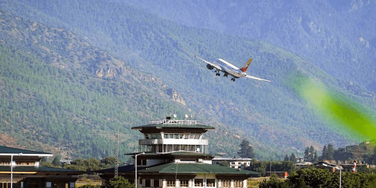 An airplane approaching the runway near a mountainous region, with an airport tower and green hills in the background, showcasing the scenic Bhutanese landscape.