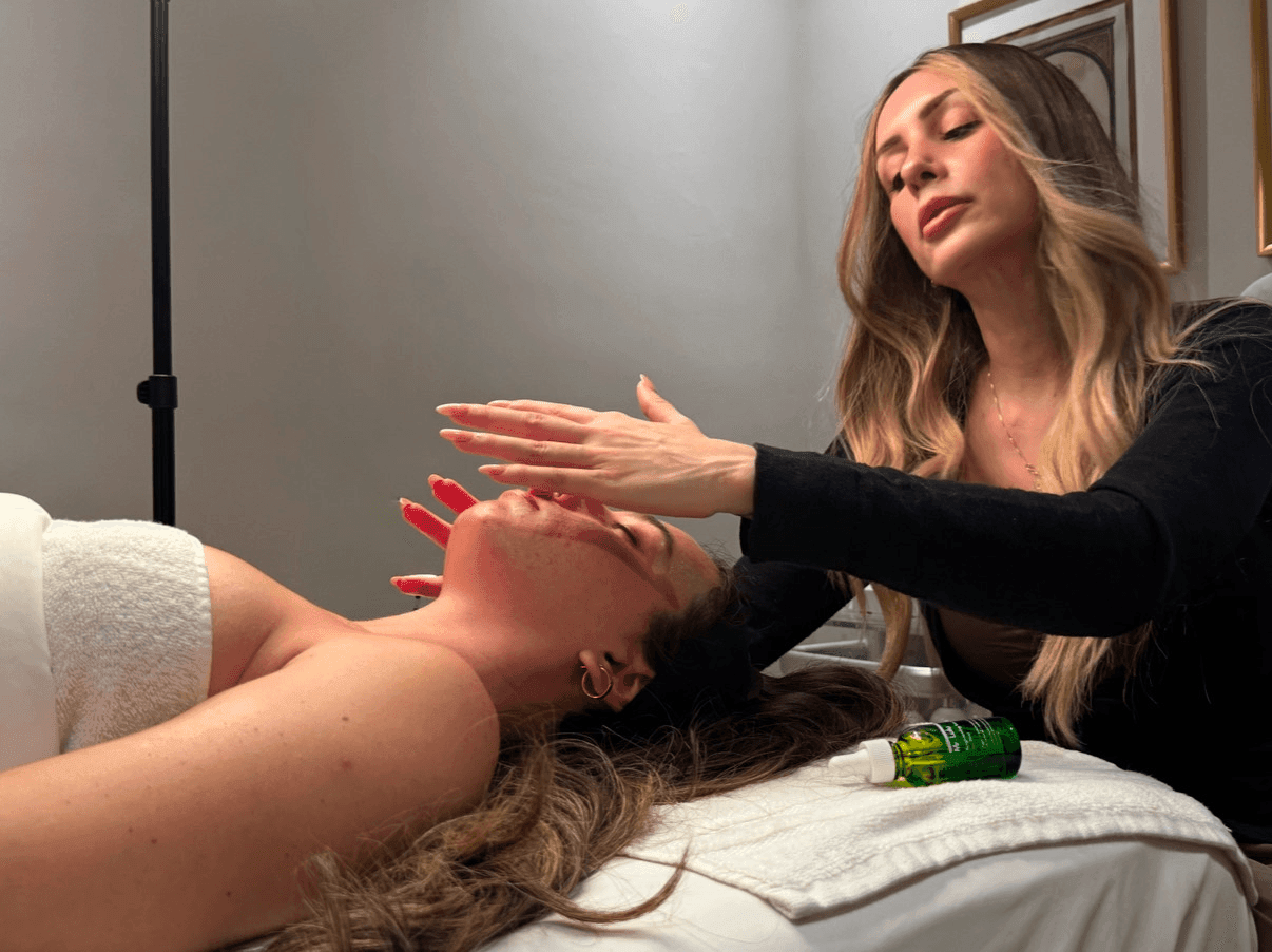 A beauty therapist applies skincare treatment to a client's face during a facial treatment session. The client, lying on a treatment bed, is receiving a facial massage, with a bottle of oil visible beside them. The environment is calm and serene, with soft lighting and minimalistic decor.