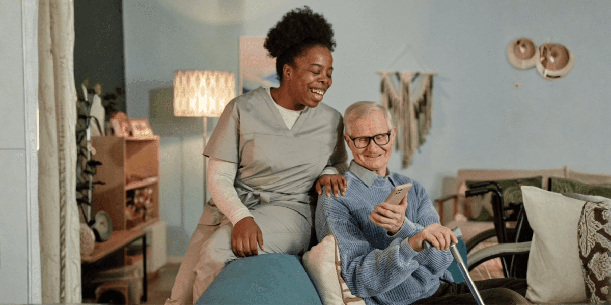 Caregiver and elderly man smiling together in a cozy living room, with the man holding a phone.