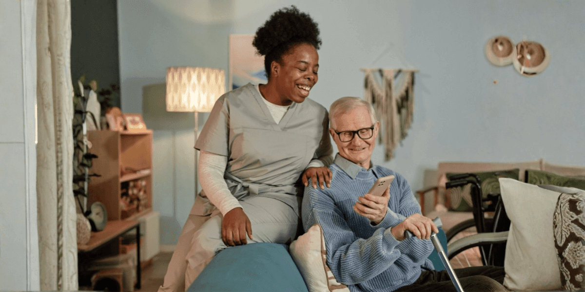 Caregiver and elderly man smiling together in a cozy living room, with the man holding a phone.
